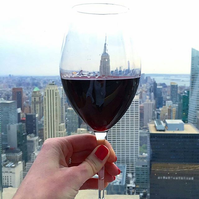 A woman's hand holding a glass of red wine in front of the NYC skyline.
