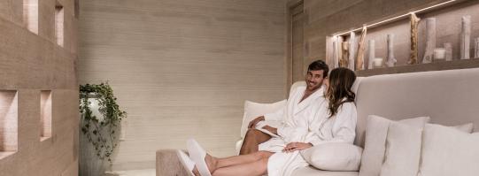 A man and woman in white robes and slippers chatting in a spa waiting area.