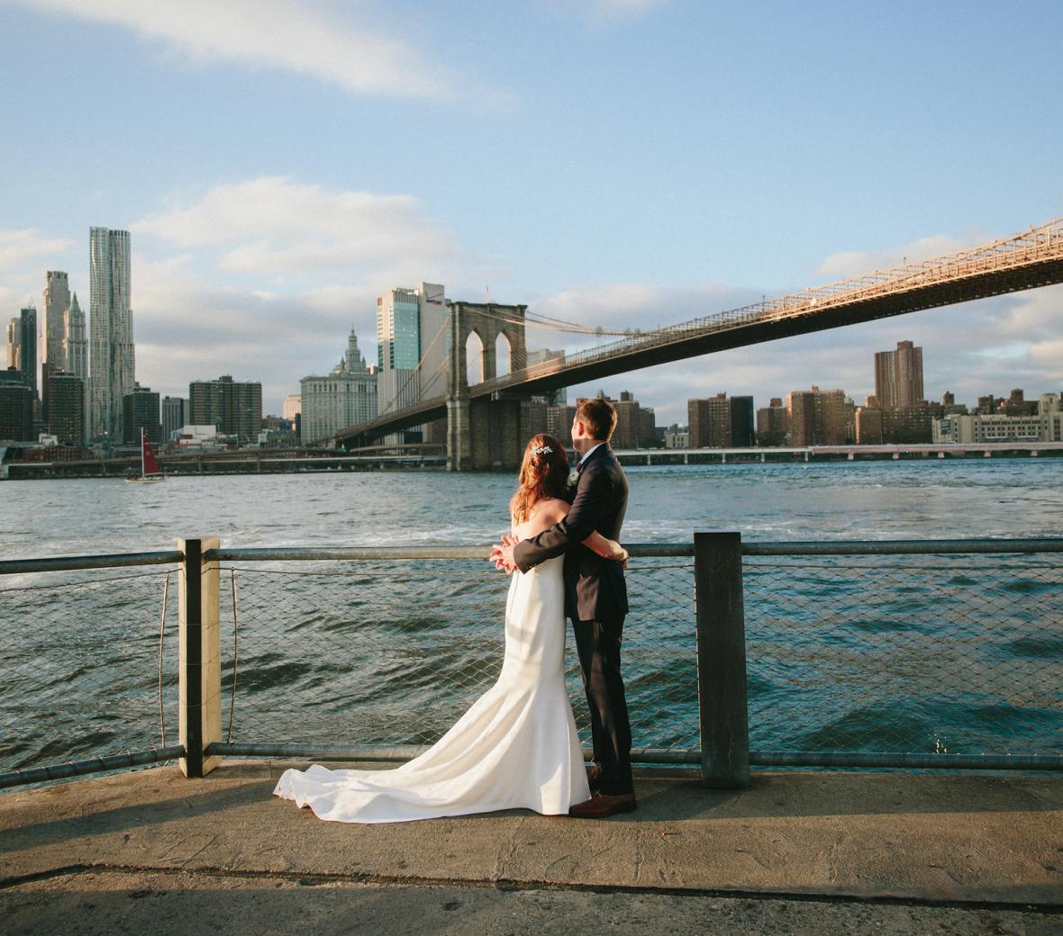 Couple stands on waterfront near brooklyn bridge in wedding attire